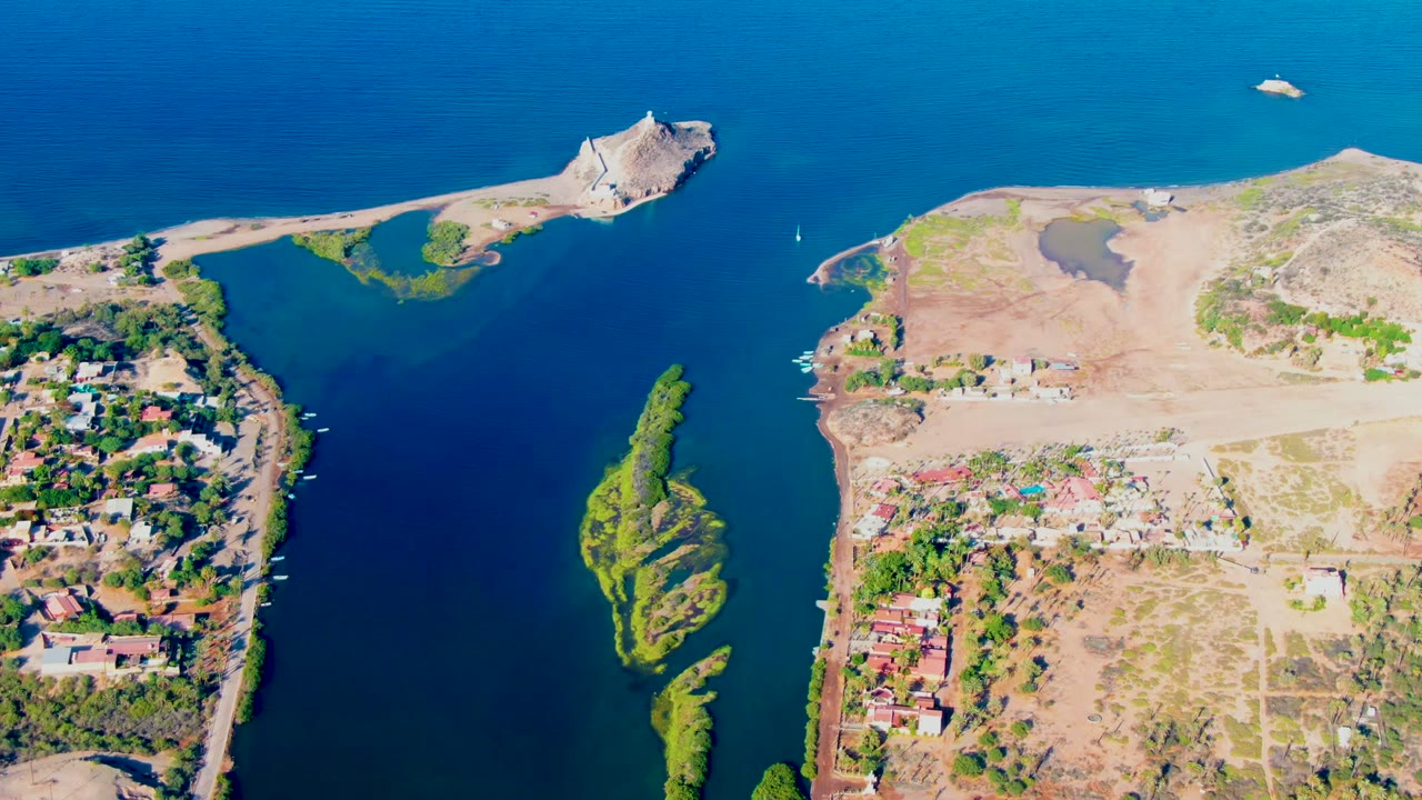 Drone-style overhead view of the channel with the sailboat, island vegetation, homes, and construction land.