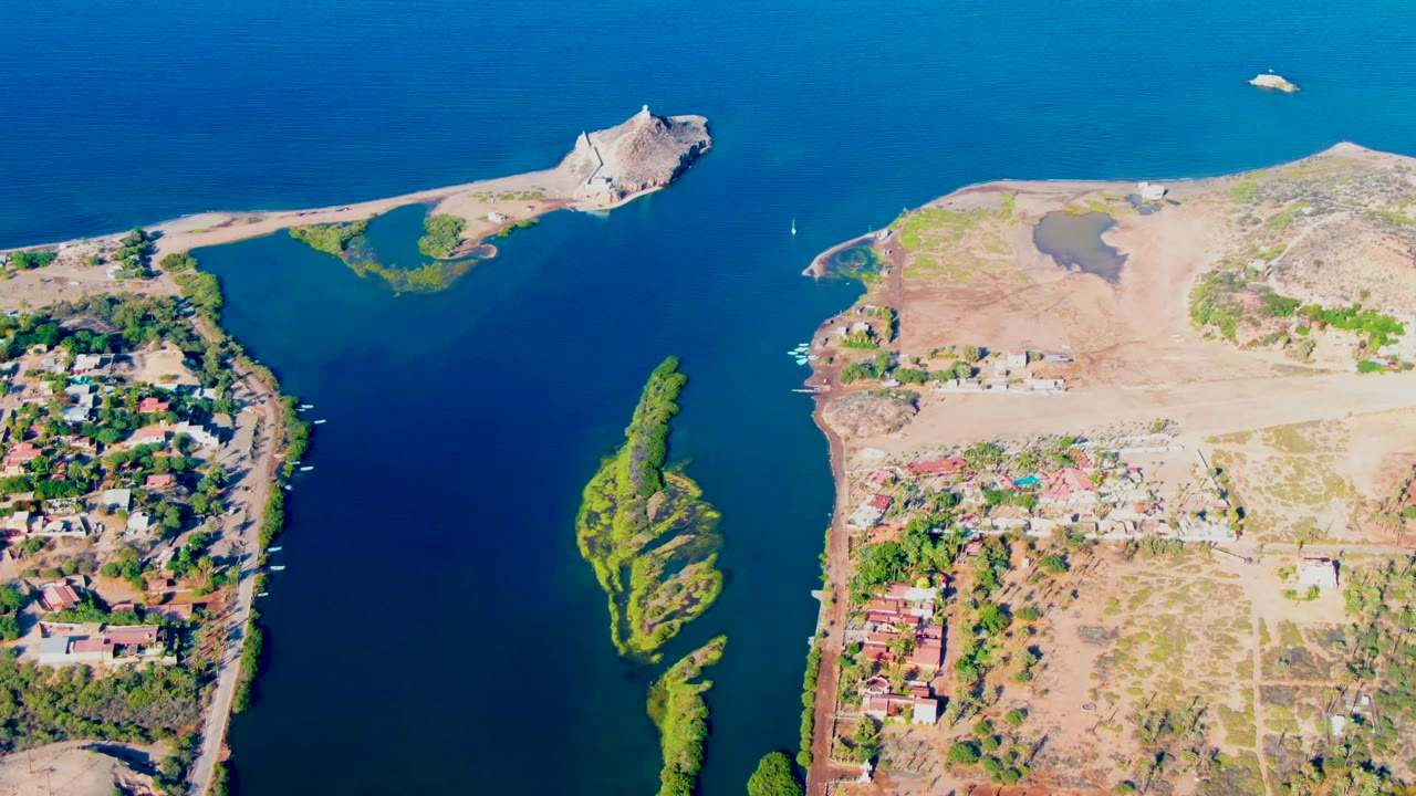 A Mulege aerial frame shows the sailboat crossing the channel between residential shoreline on the left and open dry construction land on the right, with the green island strip below and no visible people.