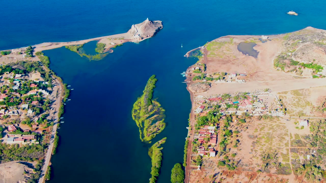 High aerial frame over Mulege, Baja California Sur, showing a blue waterway, a narrow green island, dry tan shorelines, scattered buildings, and no visible people.