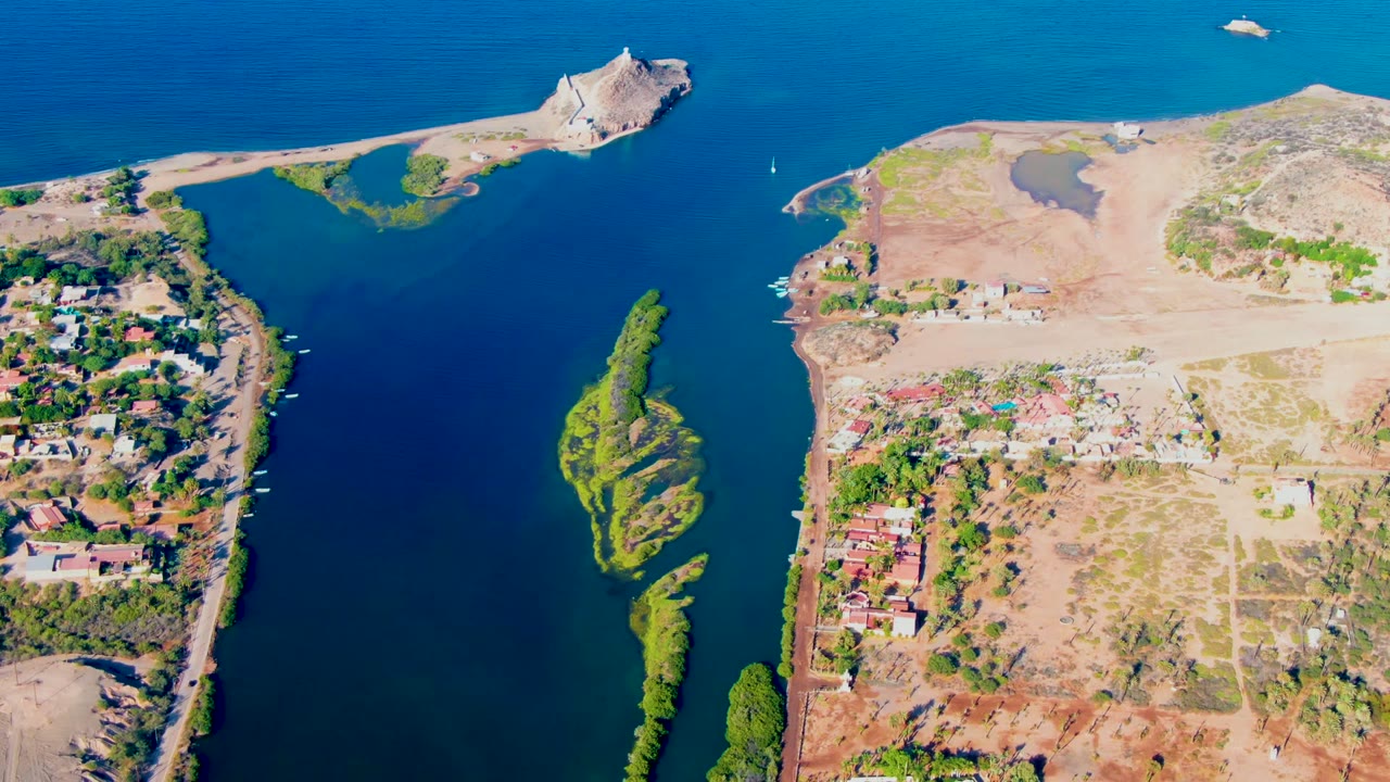 Aerial frame over Mulege, Baja California Sur, showing the boat shifted across the blue channel, the elongated green island below it, dry shore parcels, buildings, and no visible people.