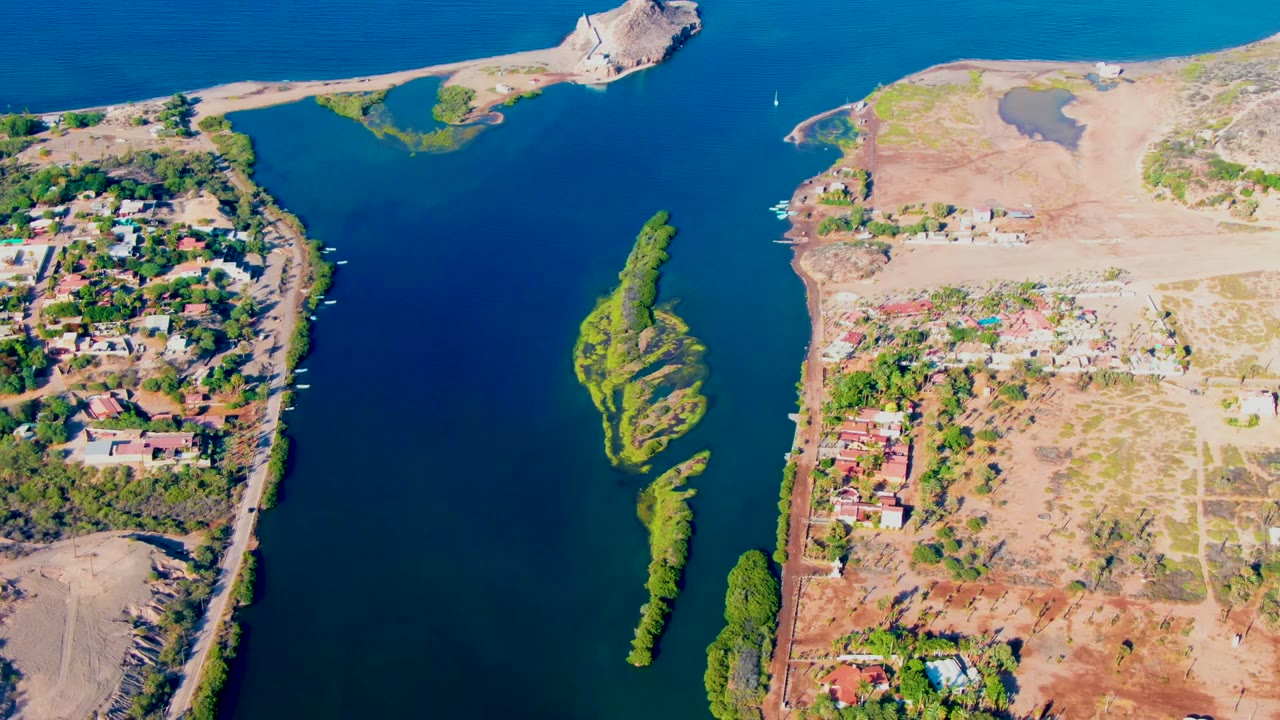 Aerial frame over Mulege, Baja California Sur, showing blue water around a narrow green island, dry shorelines, clustered homes, trees, and no visible people.