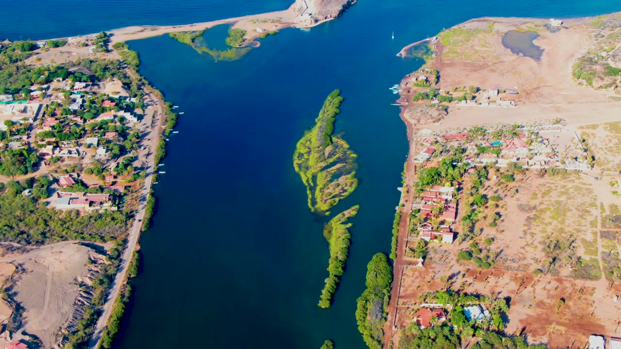 High aerial view over Mulege showing the blue channel, small green island, sandy upper shoreline, scattered homes, tree cover, and no visible people.