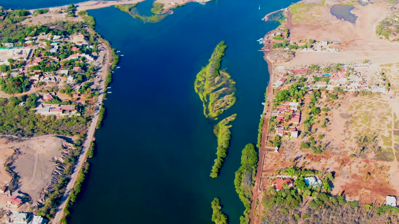 Aerial frame over Mulege, Baja California Sur, showing blue-green river water between houses on the left bank, dry open land on the right bank, tree-covered islands, and no visible people.