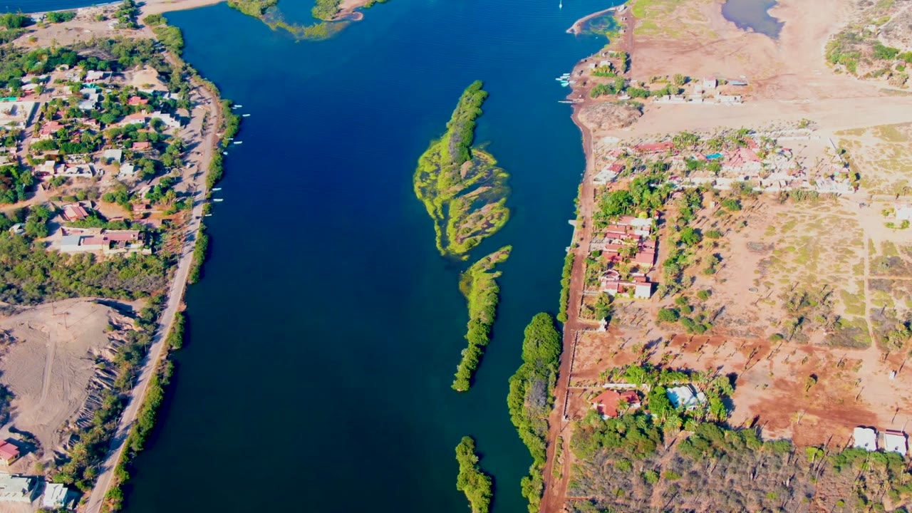 Aerial frame over Mulege, Baja California Sur, showing the river bend, clustered houses near trees, dry graded-looking land, small islands in the water, and no visible people.
