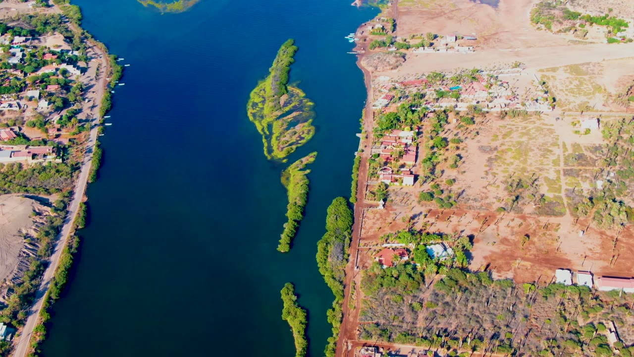 Overhead view in Mulege, Baja California Sur showing deep blue river water, narrow green islands, shoreline homes, dry open land, and no visible people at the start of the clip.