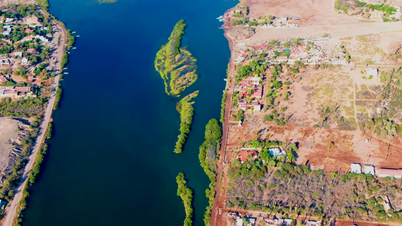 Top-down Mulege river view with elongated green islands in the water, a narrow shoreline road, tree clusters, scattered homes, open land, and no visible people.