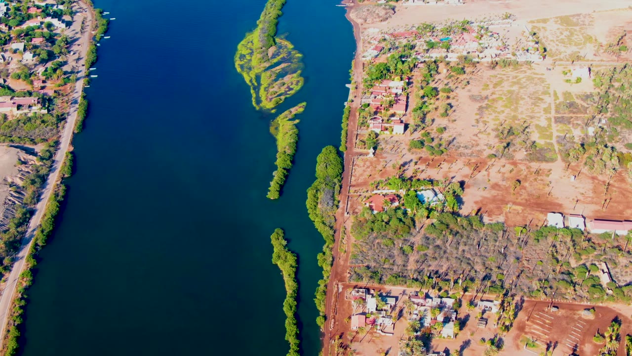Overhead view in Mulege, Baja California Sur, with deep blue river water, dry brown shoreline, roads, scattered buildings, and narrow green islands, no visible people.