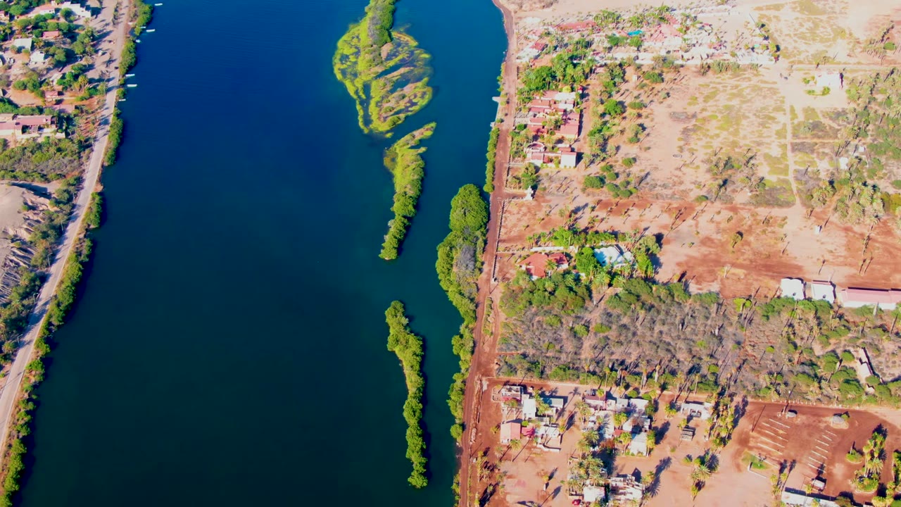 High overhead view in Mulege with dark blue water, thin green vegetated islands near the center, dry bank and developed land along the right side, no visible people.