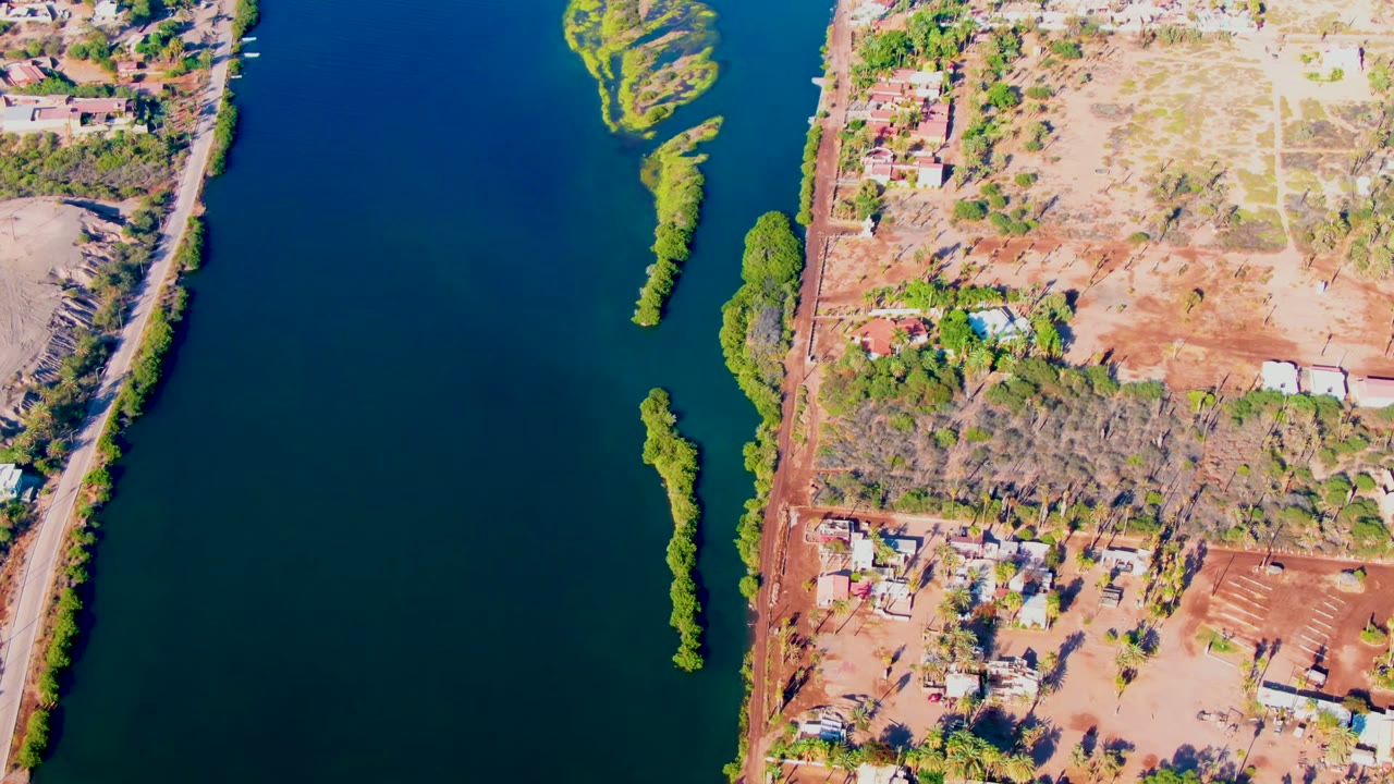 Overhead frame in Mulege, Baja California Sur showing blue river water, a narrow green island, reddish dry land, scattered buildings, trees, roads, and no visible people.