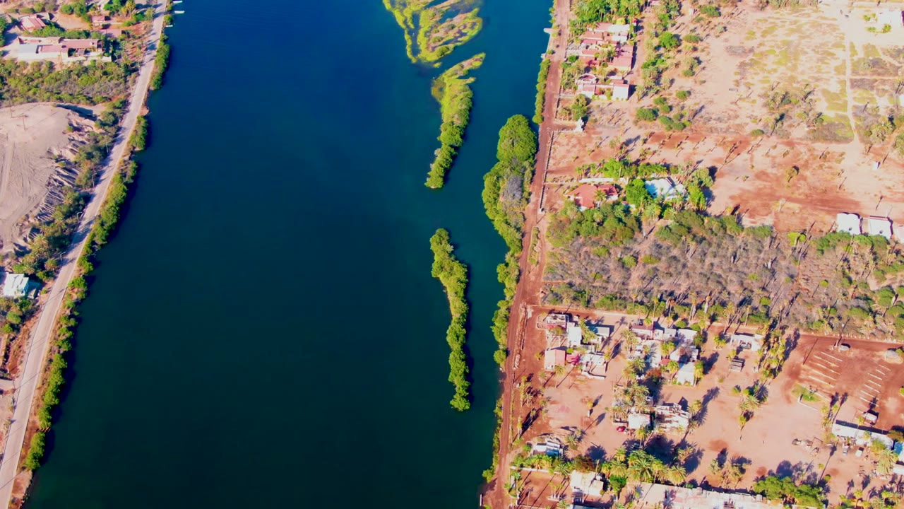 High-angle Mulege aerial showing a broad section of blue river, a narrow vegetated island, dry tan and red land, scattered riverside buildings, roads, trees, and no visible people.