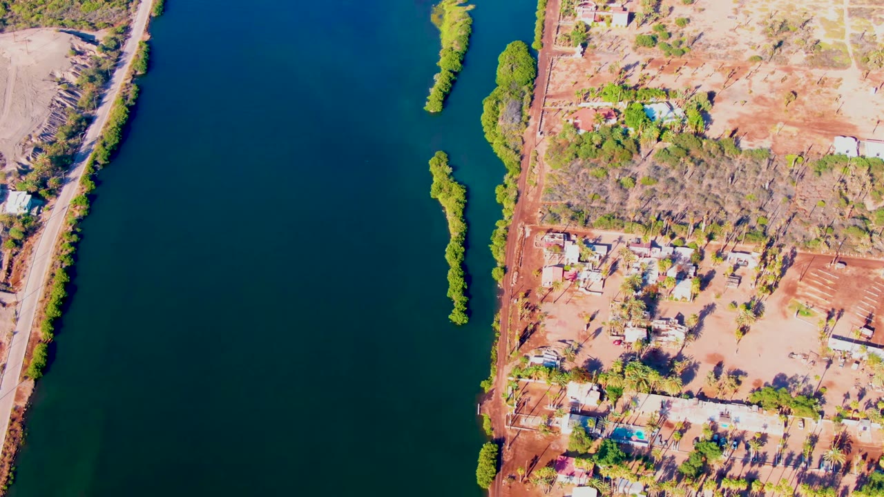 Overhead frame from Mulege, Baja California Sur showing blue-green river water, a narrow tree-covered island, dry land, dirt roads, scattered buildings, and no visible people.