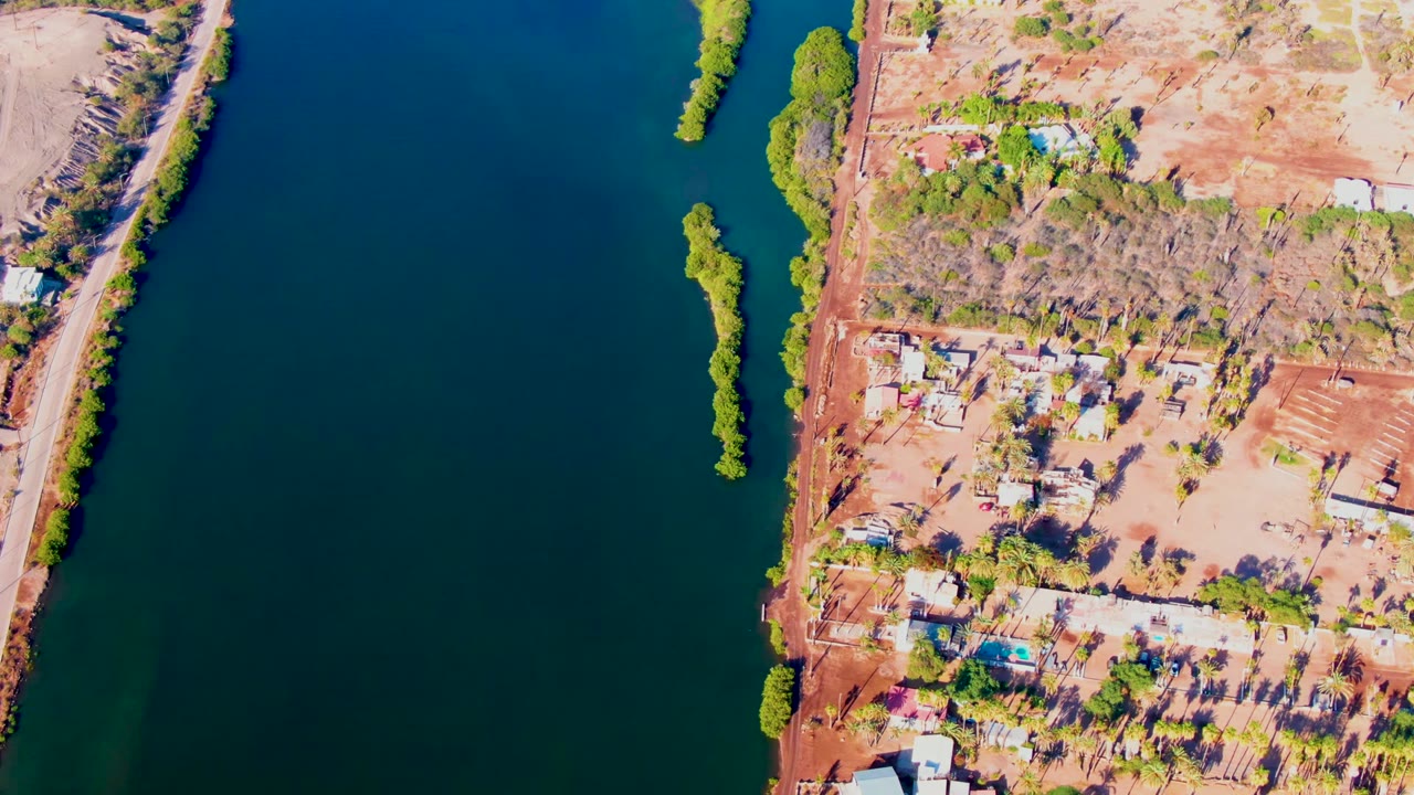 Mulege overhead frame with dark blue-green water across the left side, a narrow vegetated island in the river, dry settlement land on the right, and no visible people.