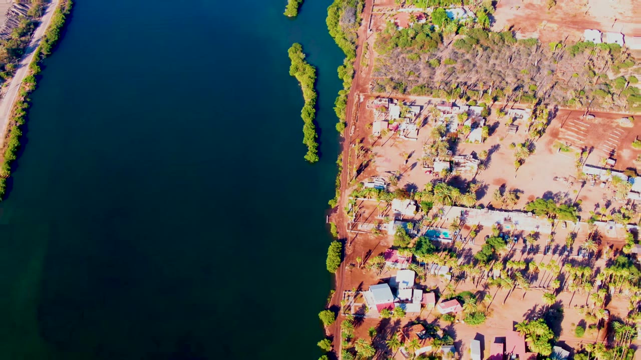 Overhead view in Mulege, Baja California Sur, showing dark river water beside reddish shoreline, scattered homes, trees, and no visible people at the opening frame.