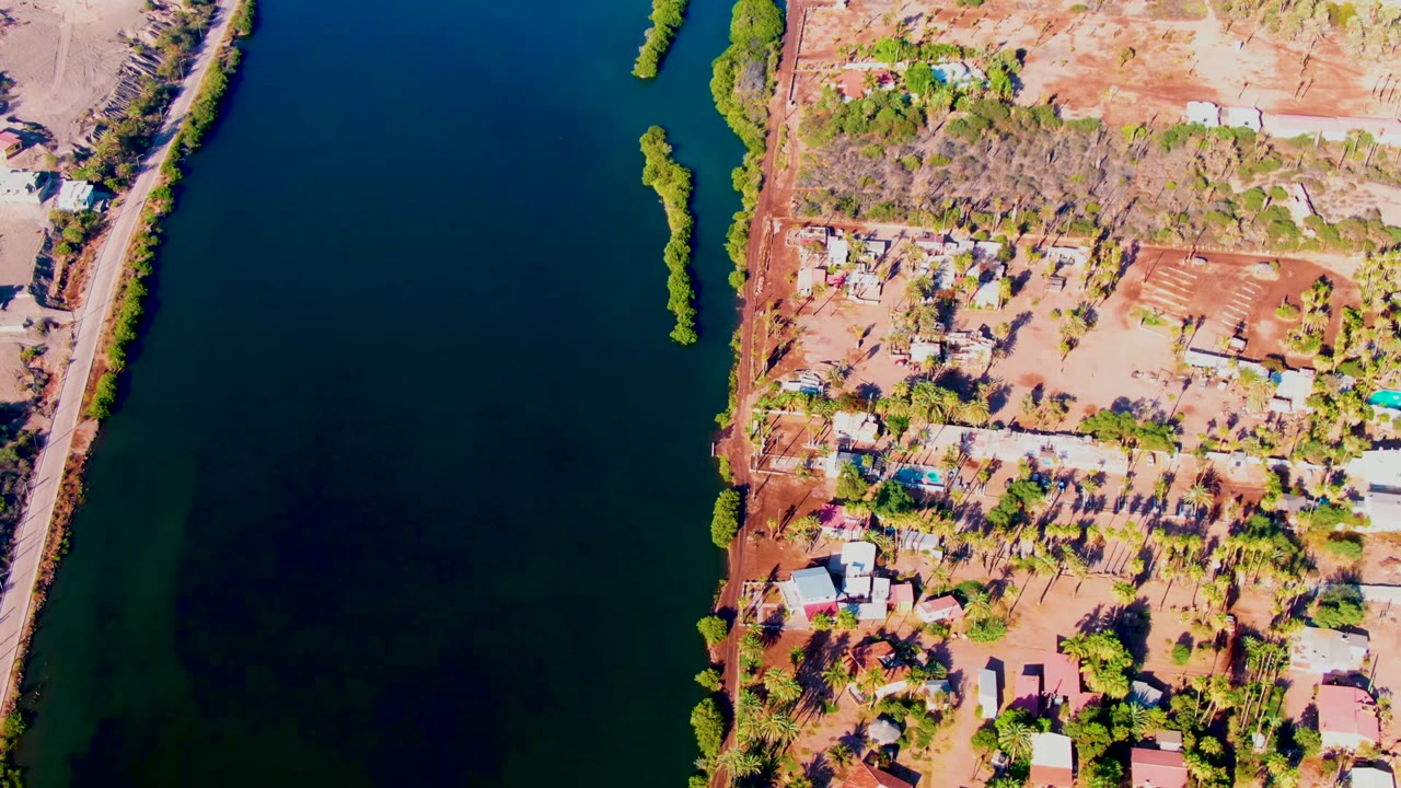 High drone view in Mulege, Baja California Sur, showing a narrow shoreline settlement between dark river water, reddish soil, tree cover, and no visible people.