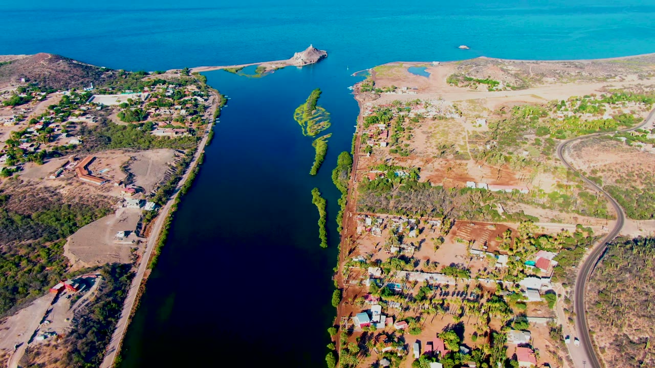Opening aerial frame over Mulege, Baja California Sur, showing a dark canal between arid landmasses, scattered buildings, roads, turquoise open water, and no visible people.