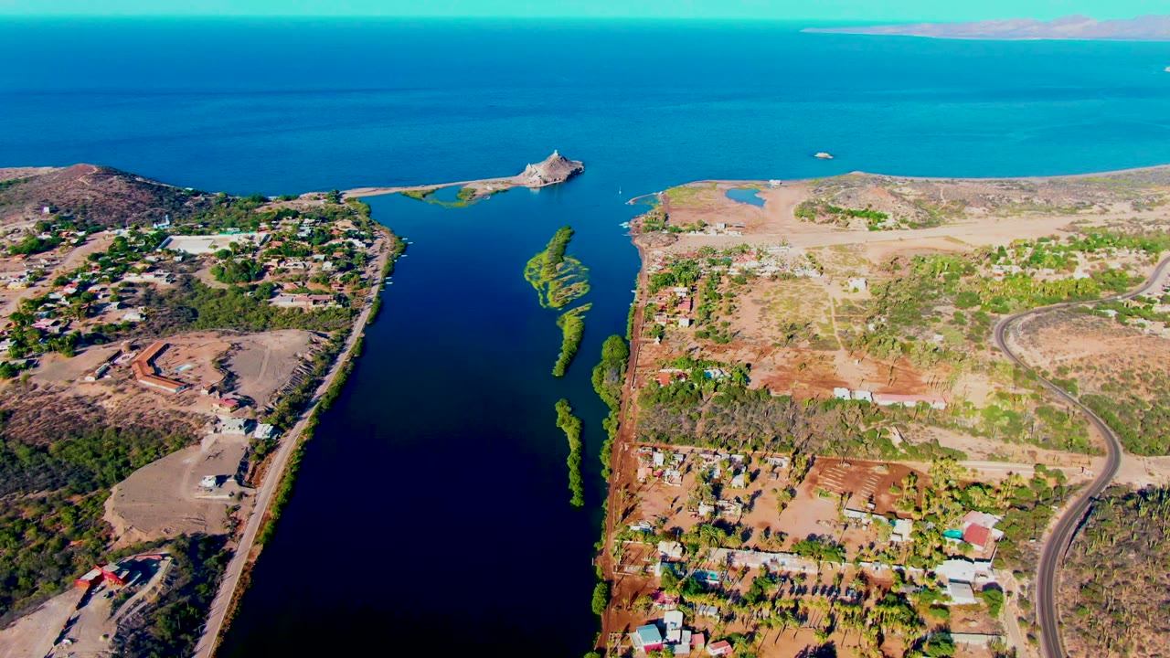 Mid-clip aerial frame in Mulege shows the dark canal, narrow green patches in the water, dry land with roads and buildings, distant open water, and no visible people.