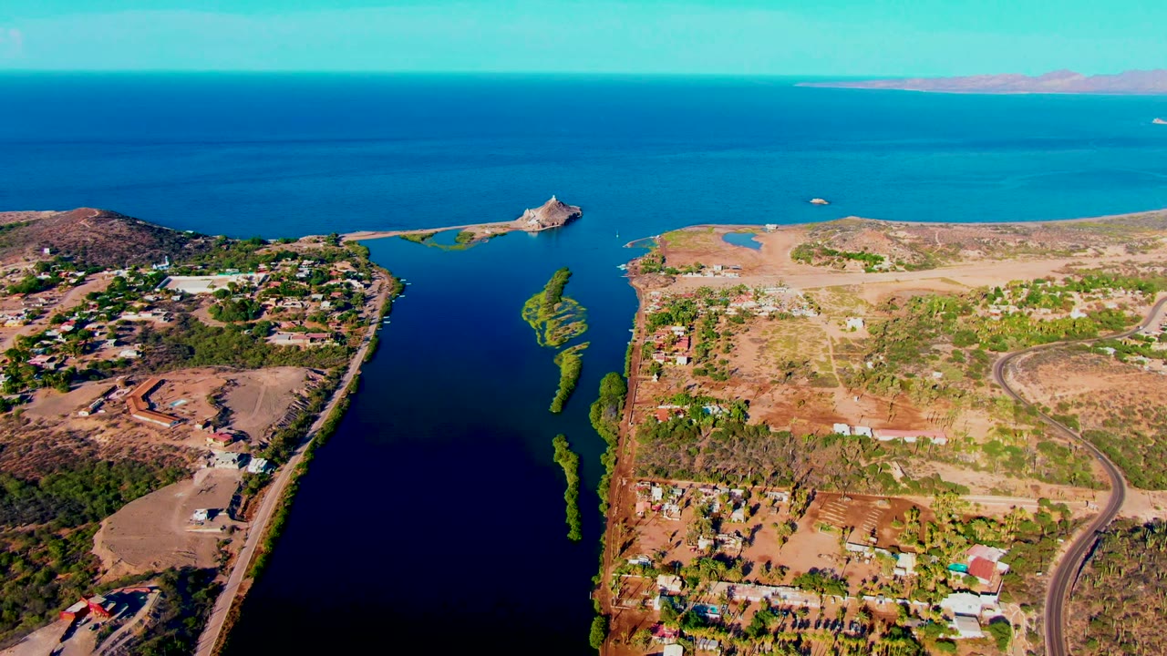 Opening frame of Mulege, Baja California Sur from above, with a dark inlet at left, dry land and roads at center, scattered houses at right, blue ocean beyond, and no visible people.