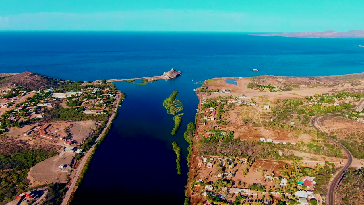 Mulege aerial frame with dark inlet water in the lower left, green patches through the channel, tan open ground, clustered houses, a small red roof, blue ocean, and no visible people.