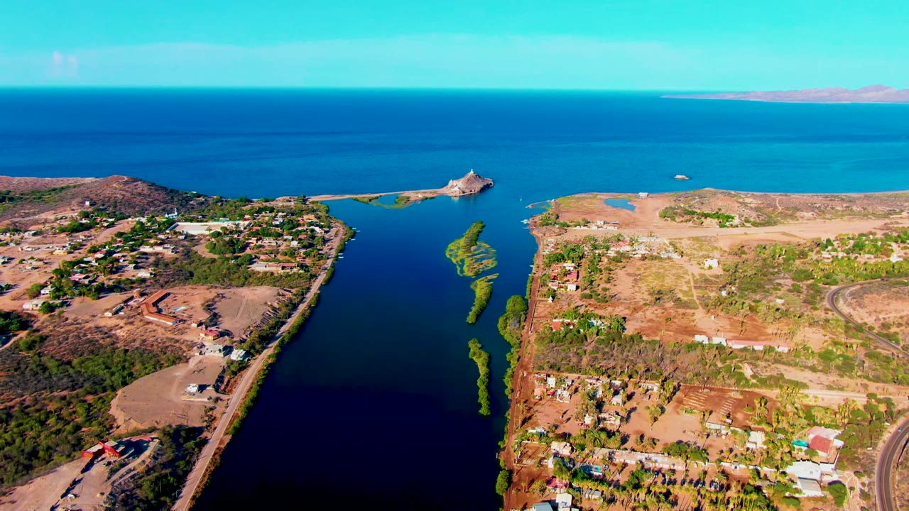 Aerial frame over Mulege, Baja California Sur, with a dark inlet between dry land and scattered buildings, the coastal road along the lower edge, blue sea beyond, and no visible people.