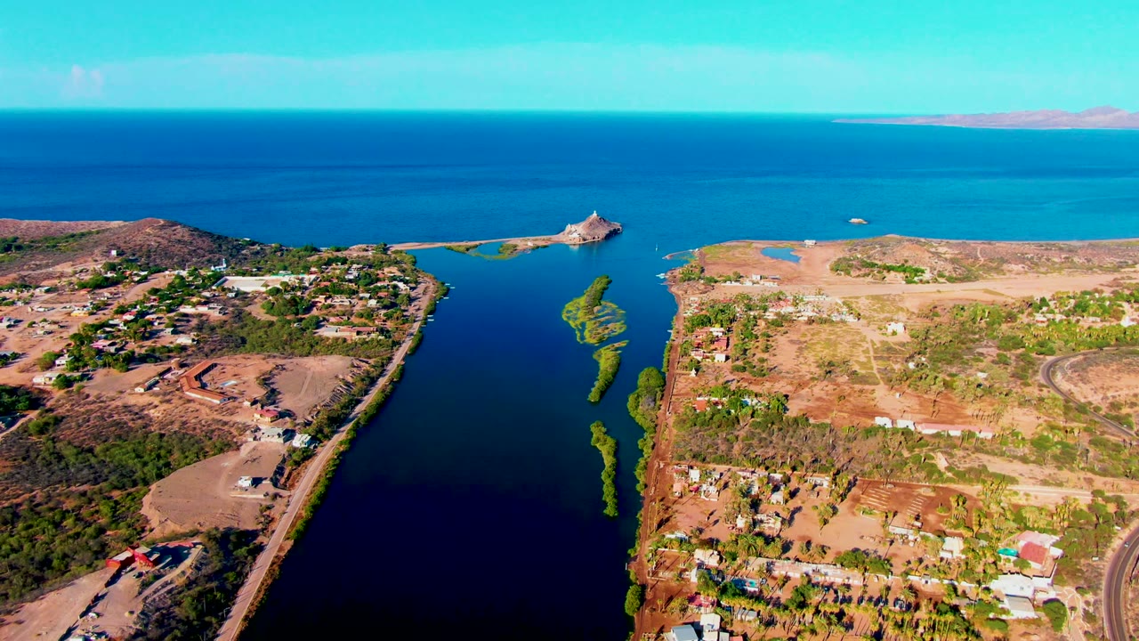 Drone view of dark inlet water beside dry terrain and a road near scattered homes.