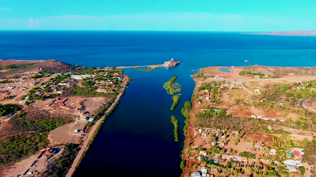 High aerial frame over Mulege with the narrow waterway running toward the sea, a road tracing the land edge, small buildings below, dry lots to the right, and no visible people.