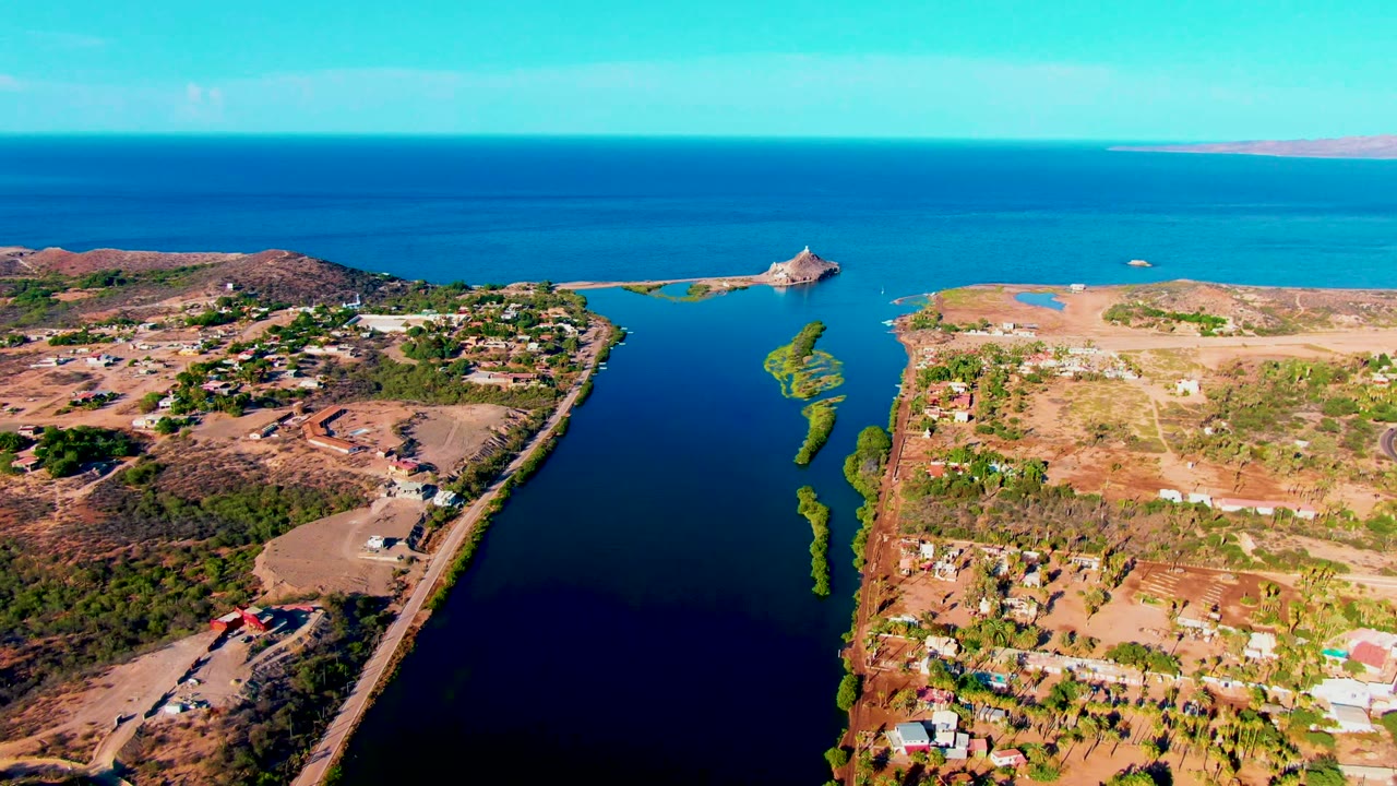 Opening aerial frame over Mulege, Baja California Sur, showing a dark canal, dry shore roads, scattered homes, turquoise sea, distant low land, and no visible people.