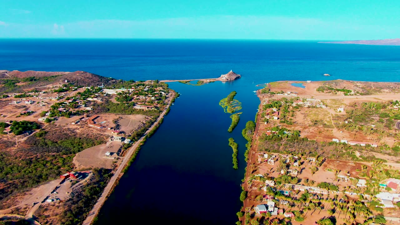 Aerial canal view with dry shoreline, homes, roads, green water plants, and open sea beyond.
