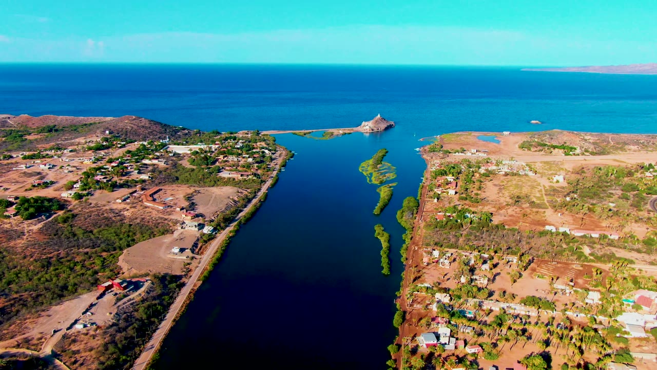 Mid-clip aerial frame in Mulege shows the dark canal flanked by tan arid land, small building clusters, a curved road, bright coastal water, and no visible people.