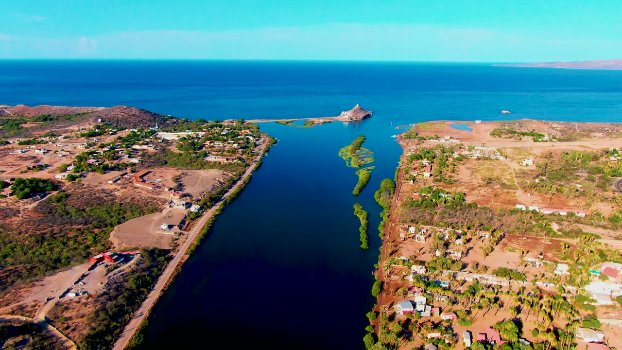 Opening aerial frame over Mulege, Baja California Sur, showing a narrow blue waterway, dry land on both sides, scattered buildings, shoreline greenery, distant open water, and no visible people.
