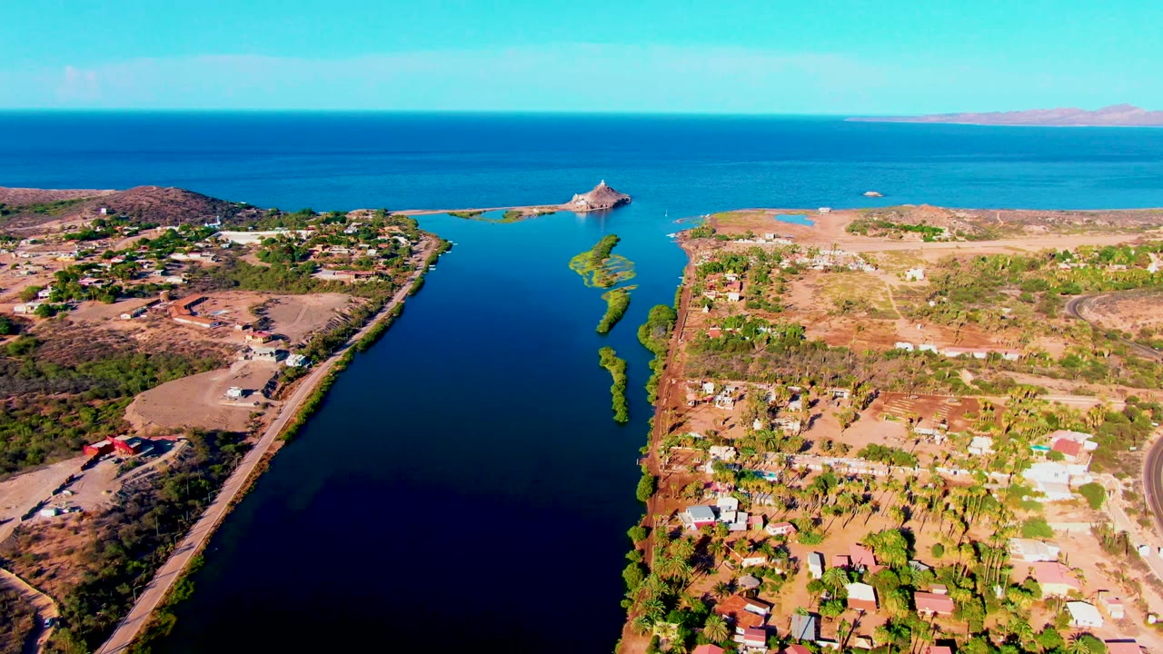 Opening aerial frame over Mulege, Baja California Sur, with a dark canal in the foreground, a road along the right bank, scattered houses, turquoise sea, low islands, and no visible people.