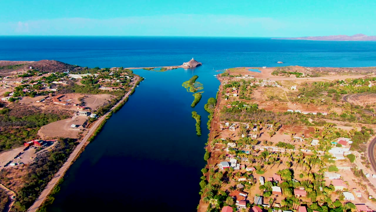 Drone view of canal water, roadside, houses, dry terrain, and distant coastline.
