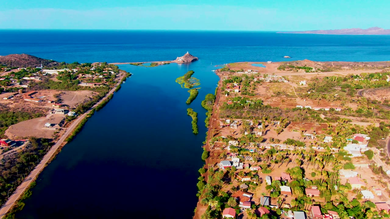 Aerial frame over Mulege, Baja California Sur, showing a narrow blue waterway, dry shoreline, scattered buildings, trees, open sea, and a distant island structure with no visible people.