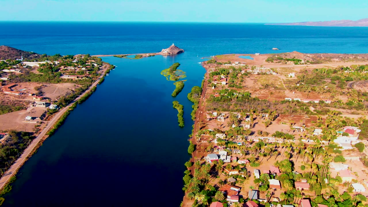 Drone view along a coastal inlet with buildings on dry land and a small island structure ahead.