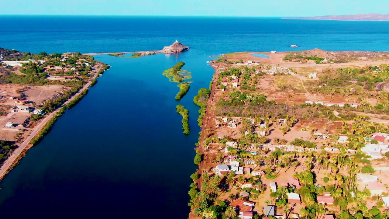 Mulege aerial frame showing calm blue water between two dry shorelines, clustered buildings near the lower edge, green vegetation strips, and the distant island structure with no visible people.