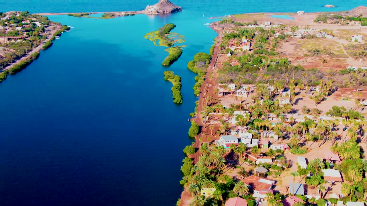 Aerial frame over Mulege, Baja California Sur showing a deep-blue inlet between dry shorelines, scattered homes, palms, turquoise ocean, a small rock island, and no visible people.