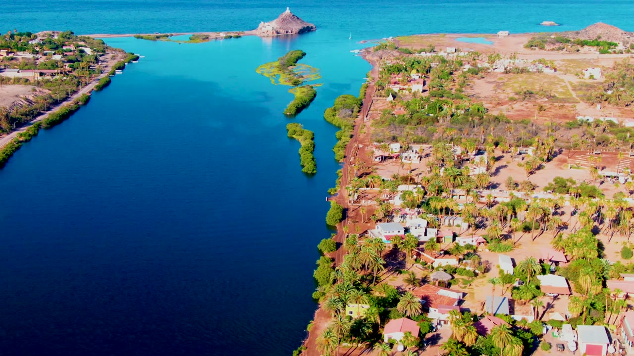 Drone view looking along an inlet beside dry land, palms, homes, and distant ocean.
