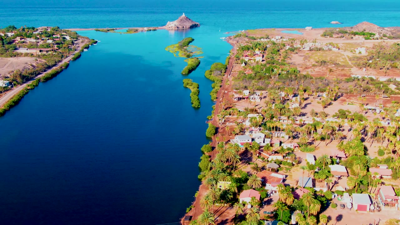 Drone aerial over Mulege, Baja California Sur showing the inlet beside a denser cluster of homes and palms on the right, dry open ground inland, blue ocean beyond, and no visible people.