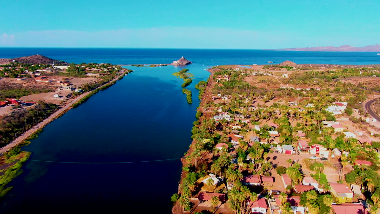 Opening frame of Mulege, Baja California Sur from above, with a dark blue canal on the left, homes and palm trees along the shore, ocean and distant hills beyond, and no visible people.
