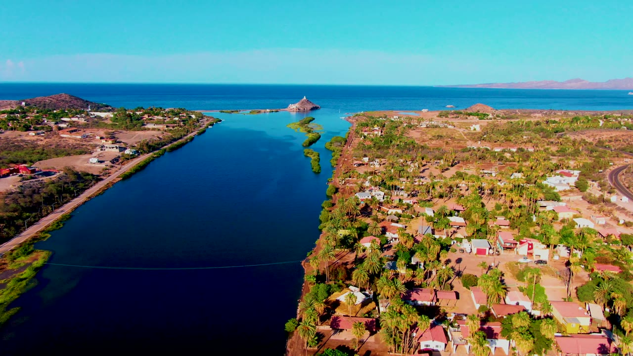 Aerial canal view with shoreline houses, palms, ocean horizon, and a small rocky islet.