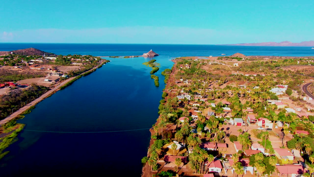 At 0:02, the Mulege canal and palm-lined homes sit below the camera, with dry ground and a curved road to the right, blue ocean across the background, and no visible people.