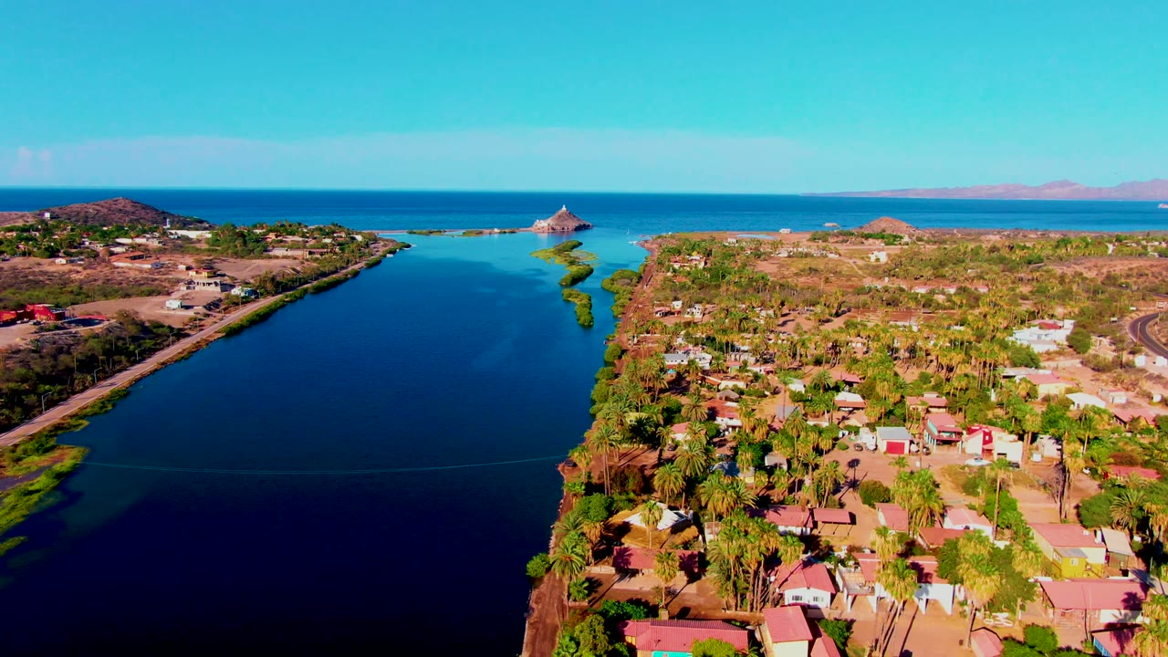 Opening aerial frame over Mulege, Baja California Sur, showing a blue canal between residential shoreline, palm trees, distant ocean, low hills, and no visible people.