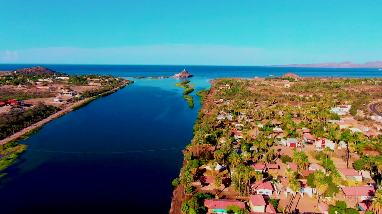 Wide drone view of a canal, waterfront houses, palms, and open ocean beyond.