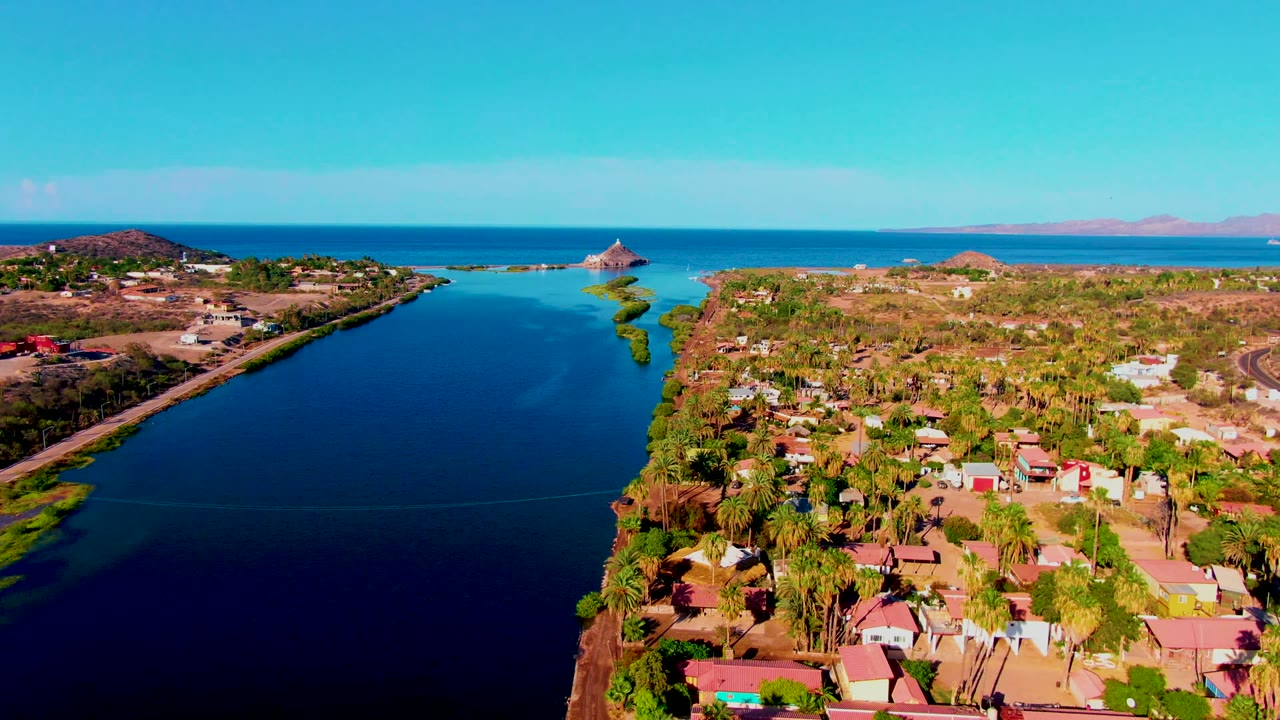 Mulege aerial frame showing a dark blue canal, a palm-filled residential area, green canal edges, the ocean beyond, and no visible people.