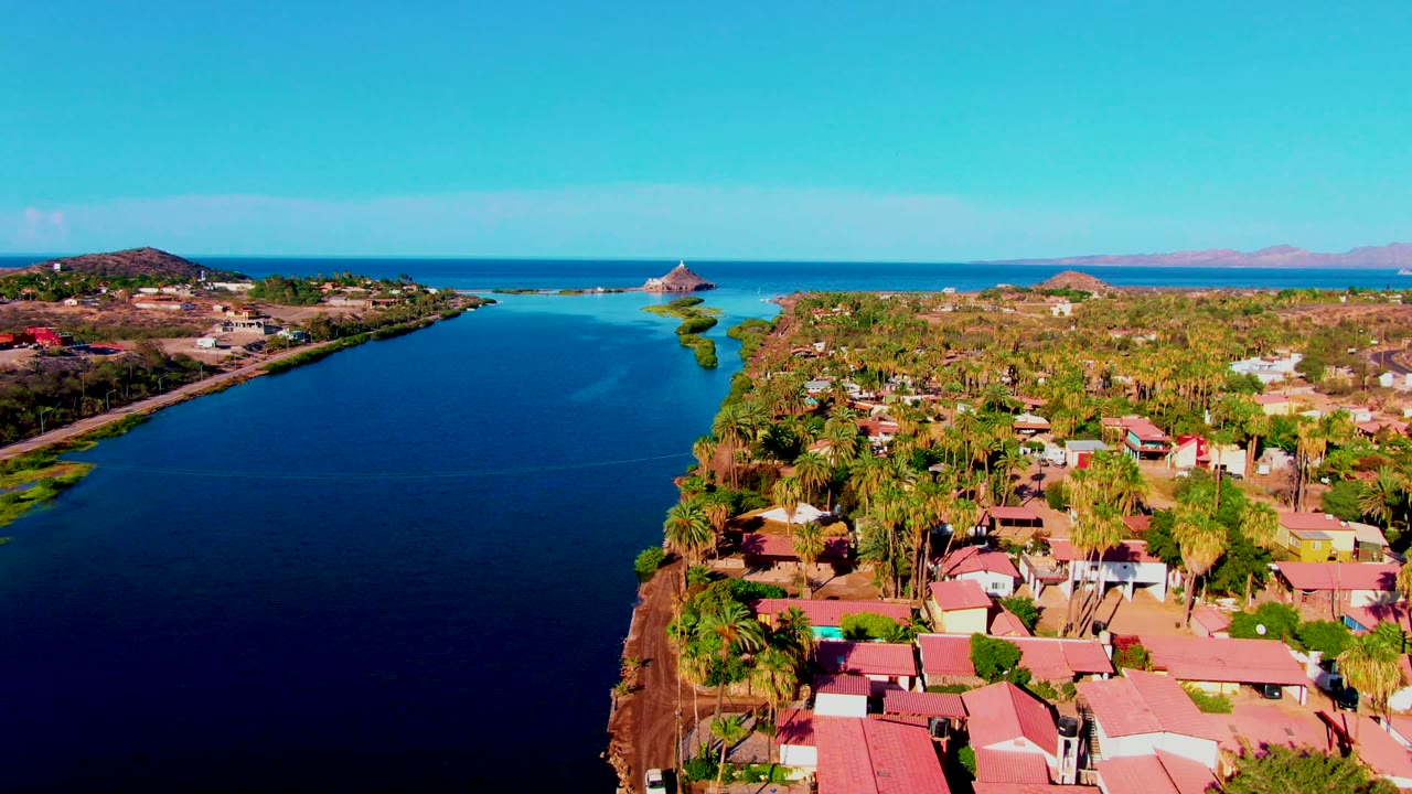 Opening aerial frame over Mulege, Baja California Sur, with dark blue canal water beside red-roofed homes, palms, open ocean, and no visible people.