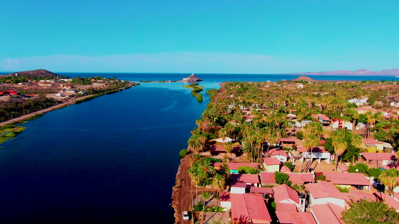 Early drone frame in Mulege showing the canal widening through the left side of the image, homes and palms along the right bank, ocean beyond, and no visible people.