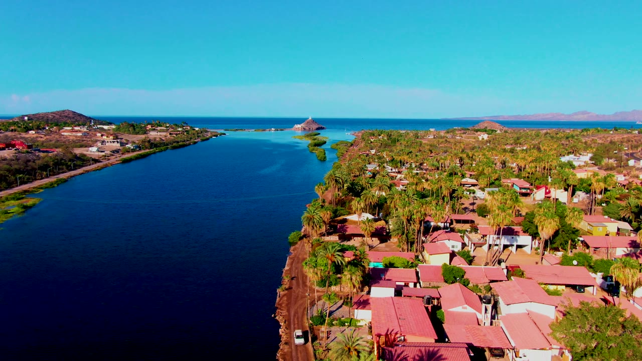 Mid-clip aerial over Mulege with calm blue water on the left, a dense cluster of palms and red-roofed houses on the right, distant ocean, and no visible people.