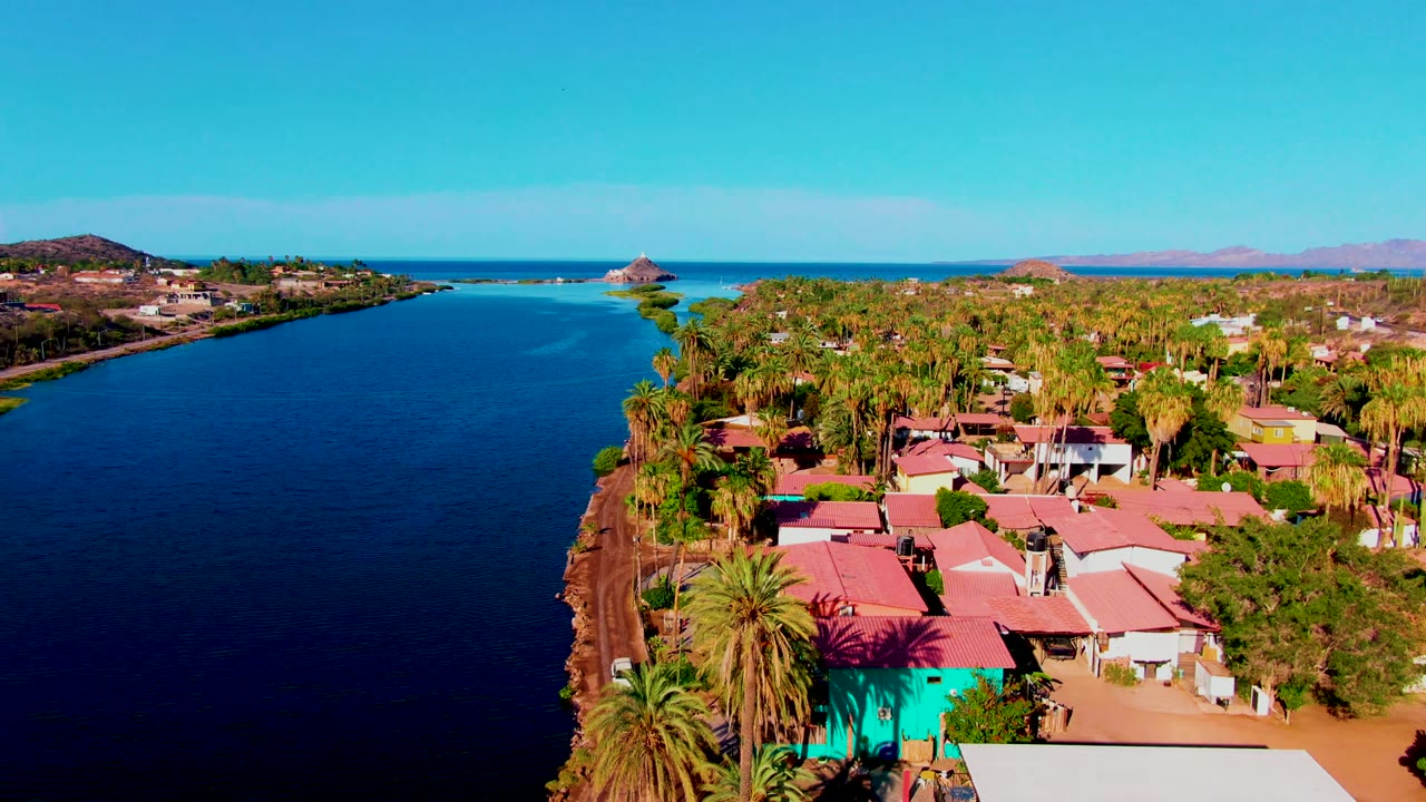 Opening aerial frame over Mulege, Baja California Sur, showing a broad blue waterway, red-roofed homes, palms, low hills, ocean horizon, and no visible people.