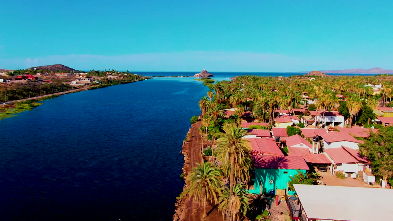 High oblique aerial frame of Mulege, Baja California Sur, with dark blue water at left, a palm cluster and red-roofed residential buildings at right, and no visible people.