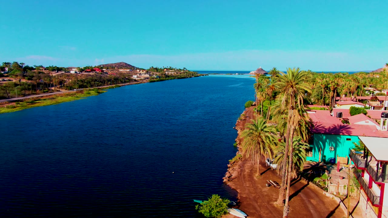Aerial view over a wide river in Mulege, Baja California Sur, with a palm-lined dirt bank, turquoise building at the right edge, distant houses, clear blue sky, and no visible people.