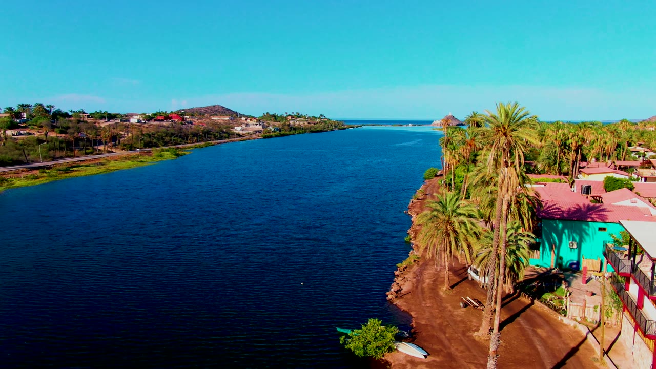 Riverbank with palms, turquoise building, dirt path, and distant houses under blue sky.